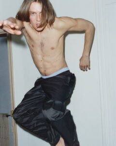 A person with long hair is captured mid-movement, appearing to leap or jump. Their arms are extended, emphasizing dynamic motion. They are shirtless, wearing dark, loose-fitting pants with a light waistband. The background shows a simple interior space with a door slightly ajar and a glimpse of a radiator, suggesting an indoor setting.