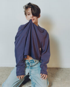 A person is kneeling on a carpeted floor, wearing a loose-fitting purple shirt and light blue jeans. The shirt is being playfully held in their mouth. Their hair is styled with a side part and partially covering the face. The background is simple, with a plain white wall, creating a casual and relaxed atmosphere.