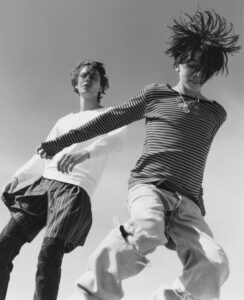 Two individuals are dynamic and expressive in their movements, set against a clear sky. One wears a long-sleeve striped shirt and distressed jeans, hair flying wildly. The other sports a white shirt over layered shorts, gazing forward with tousled hair. The scene captures a sense of youthful energy and freedom, emphasizing the carefree spirit of youth.