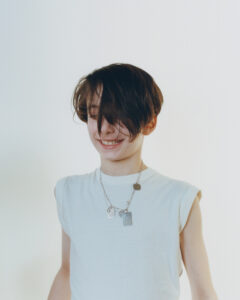 A person with dark, medium-length hair smiles candidly, eyes slightly hidden by their hair. They wear a sleeveless, light-colored shirt and a silver necklace with pendants and tags. The background is plain and light in color, creating a soft, minimalist atmosphere. The relaxed expression and casual attire give a sense of youthful energy and simplicity.