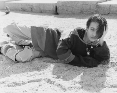 A person with short hair is lying on sandy ground, wearing a striped sweater and casual pants. They rest their head on one hand, looking towards the viewer with a thoughtful expression. The scene is in grayscale, creating a timeless, nostalgic feel. In the background, there are steps and a small, blurry figure seated in the distance.