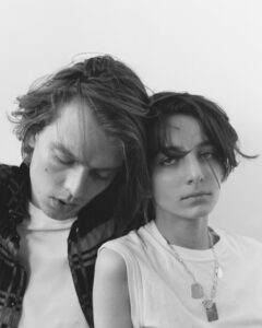 Two people appear close together, exuding a relaxed and introspective vibe. One person wears a checkered shirt over a light top, their eyes closed slightly. The other person has short, tousled hair, gazing thoughtfully at the camera while wearing a sleeveless top and a layered necklace. The monochrome tones lend a timeless, classic feel.