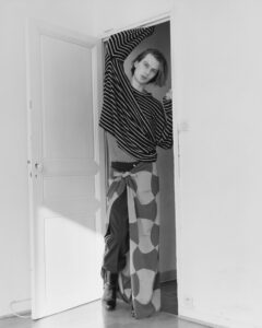 A person stands in a doorway, leaning casually. They wear a striped sweater and unique wide-legged pants with a flowing, patterned scarf-like overlay. One arm extends upward, touching the door frame. The minimalistic setting features a partially open door, soft light casting subtle shadows on the floor, and a plain white wall enhances the serene atmosphere.