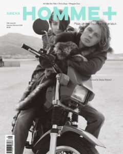 Two individuals pose on a motorcycle, with one seated behind the other, both wearing fur-collared jackets. Their expressions are calm and introspective. The setting appears to be outdoors, with distant mountains and a blurred background, suggesting a casual yet stylish atmosphere. The composition evokes a sense of adventure and fashion-forward attitude.