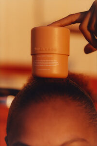 A cylindrical container with a matte finish is balanced on top of a person's head. A hand with fingers slightly bent is poised next to it, adding a playful elegance to the scene. The lighting is warm, casting a cozy glow over the objects, and the background features soft, blurred colors, enhancing the overall aesthetic.