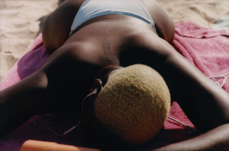 A person with short, light-colored hair is lying face down on a bright pink towel, wearing white swimwear. Earphones dangle near their shoulders, suggesting a moment of relaxation at the beach. The smooth sand surrounds the towel, reflecting a sunlit, tranquil ambiance. A nearby sunscreen bottle adds a touch of practicality to this sunbathing scene.
