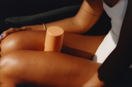 A person with warm, sunlit skin is seated, partially visible. A tan, cylindrical object rests on their thighs. The ambiance suggests relaxation, with soft lighting highlighting the skin's texture. A white outfit is worn, and a simple bracelet adorns their wrist. Subtle tattoos are visible on the arm, adding personal detail to the serene scene.