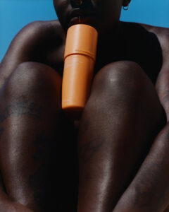 A person with tattoos sits under a clear blue sky, with knees drawn up. An orange cylindrical object is held between the lips and rests on the knees. The person has a nose ring and small hoop earrings. The close-up emphasizes the rich texture of the skin and the serene, sunlit atmosphere.