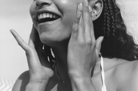 A person with braided hair is smiling joyfully. Their hands touch their face gently, applying or adjusting something on their skin. The close-up provides a view of their earrings and smooth texture of their skin. Bright lighting highlights their cheerful expression and serene moment, conveying a sense of happiness and relaxation. The style is monochromatic.