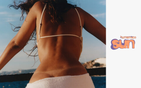 A woman with long, wavy hair stands by a railing, wearing a white bikini top. Her back is facing the viewer, showcasing a toned physique. She leans slightly forward against a towel draped behind her, with an expansive view of blue sky, calm water, and distant buildings on the horizon under a sunny day.