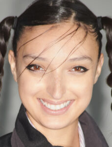 A person beams with a joyful smile, showcasing expressive eyes accented by dark eyeliner. Their hair is styled in two braided pigtails, with several wisps sweeping across the forehead. Wearing a black jacket over a light top, their cheerful demeanor radiates warmth and friendliness, exuding a lively and approachable presence.