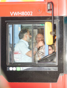 A bus driver's cabin viewed from the side shows the driver engaged in conversation with a passenger. The door's glass reflects the interior and exterior light, revealing additional passengers. The cabin's technological controls and features are visible, including side mirrors and a control panel, adding detail to the scene. Bright sunlight highlights the red exterior.