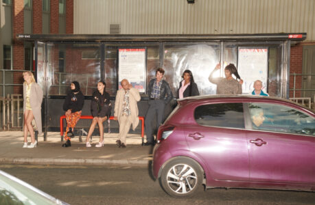A group of people stand and sit under a sheltered bus stop. They wear a variety of casual clothing, expressing individual styles. A small purple car is parked on the street in front of them. The backdrop features a brick and metal building with windows. The atmosphere appears relaxed as they wait together.
