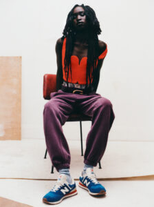 A person with long braided hair sits on a red chair against a plain background. They wear a bright orange top and purple pants with elastic cuffs. Blue and white sneakers complete the look. The setting is minimalist, with neutral tones on the floor and walls, enhancing the vibrancy of the outfit and casual, relaxed posture.