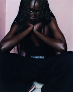 A person sits with legs apart, facing forward, resting their chin on clasped hands. Dark braided hair frames the face, and the expression is serious. The light reveals glistening skin and dark clothing, with hints of colorful footwear peeking beneath. The background is a muted pink, adding contrast to the overall composition.