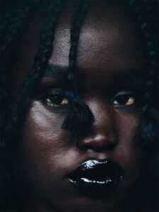 The portrait features an up-close view of a person's face with deep, rich skin tone and braided hair. The expression appears contemplative, and the lighting emphasizes the glossy, dark lips. Shadows play across the features, enhancing the intense and captivating visual effect. The braids have small sections hanging over the forehead, contributing to the overall texture and allure.