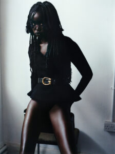 A person with long braided hair sits on a chair in a minimalist setting. They wear a stylish black outfit highlighted by a prominent belt with a gold buckle. The pose is confident and relaxed, with hands resting on either side. Soft lighting casts subtle shadows, accentuating the smooth, reflective texture of their attire.