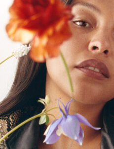 A woman gazes softly, partially obscured by a vibrant bouquet of red, white, and purple blooms. Her long, dark hair frames her face, and delicate floral details add a dreamy, ethereal quality. The composition balances warmth and beauty, highlighting the contrast between the softness of the flowers and her serene expression, creating a captivating visual harmony.