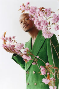 A person stands gracefully amid pink blossoms, gazing softly. They wear a vibrant green, double-breasted blazer that adds rich contrast to the delicate flowers. The simplicity of the background enhances the striking combination of colors and textures, creating an elegant and serene atmosphere. The composition emphasizes beauty and harmony between nature and fashion.