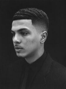 A black and white profile portrait captures a person with a neat, short haircut and a faint moustache. The individual wears a dark suit, and their expression is serious and contemplative. The background is uniformly dark, highlighting the subject's facial features and the subtle textures of their attire. The overall mood conveys elegance and introspection.