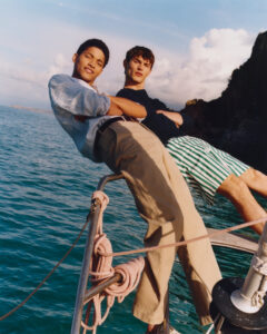 Two individuals appear relaxed on the bow of a boat, leaning back with arms folded. They are set against a backdrop of clear blue water and a distant rocky coastline under a sky filled with fluffy clouds. One wears a striped shirt and beige pants, while the other sports a dark top and striped shorts.