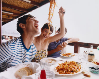 A lively gathering features three people enjoying a meal on a covered outdoor patio. The focus is on a vibrant exchange with pasta, as one person playfully holds a forkful aloft. The table is adorned with plates of spaghetti, bread, and drinks. The atmosphere is cheerful and relaxed, with an inviting view beyond the railing.