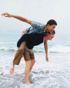 Two young people are having fun at the beach, with one giving the other a piggyback ride. Both are laughing and appear joyous, surrounded by gentle waves lapping at their legs. The sky is overcast, adding a soft, muted tone to the scene. Their casual attire suggests a carefree, playful moment by the ocean.