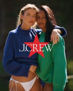 Two people stand closely together outdoors, wrapped in oversized knit sweaters in contrasting jewel tones. One rests an arm across the other's shoulders, creating a sense of intimacy, trust, and ease. Soft sunlight illuminates their faces while the blurred natural backdrop keeps attention on their connection. The styling balances comfort and sophistication, evoking warmth, companionship, and effortless contemporary elegance.