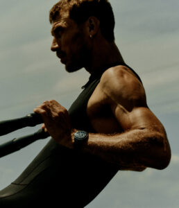A muscular individual is captured in a dynamic pose, rowing a boat. The figure is wearing a black sleeveless garment, emphasizing their toned physique. A wristwatch catches the light, contrasting against the sunlit, slightly hazy backdrop. Their determined expression and focused gaze contribute to a sense of strength and concentration in an outdoor setting.