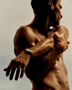 A muscular individual is captured from the side, performing a stretch that highlights their toned physique. Their left arm reaches across the body to grasp the right shoulder, revealing defined muscles and glistening skin. A prominent watch adorns the left wrist, contrasting against the skin. Soft light enhances the contours of their body against a neutral background.