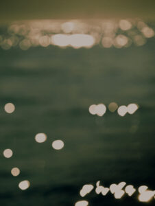 A softly blurred scene features undulating water, bathed in warm, golden sunlight. The surface is sprinkled with glistening highlights resembling twinkling stars. This dreamy, abstract representation evokes a serene, tranquil ambiance, inviting contemplation. The gentle bokeh effect enhances the play of light, making the scene feel peaceful and ethereal, reminiscent of a quiet afternoon by the sea.