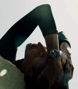 A person with closed eyes leans back, hands gracefully cradling their head. They wear a dark, long-sleeved top, and a stylish watch adorns one wrist, catching the light. The pose exudes calm and introspection, enhanced by soft focus and gentle lighting. Subtle textures and shadows create depth, while a serene atmosphere envelops the scene.