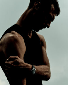 A muscular person, seen in profile against a muted background, gazes downward. Their arm is bent, with the opposite hand resting on the bicep. The individual wears a sleeveless black top, and a watch adorns their wrist. Shadows accentuate the defined muscles, creating a dramatic and contemplative atmosphere, suggesting strength and introspection.