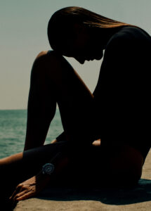 A silhouette of a person sits on a surface, knees bent and head resting forward. They wear a form-fitting outfit, with a watch on one wrist. The background features a calm sea meeting a clear sky, creating a serene atmosphere. The lighting casts dramatic shadows, emphasizing the contours and posture in an introspective pose.