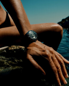 A tanned arm and hand rest on a rocky surface beside clear water, highlighting a sleek, dark wristwatch. The scene captures the essence of summer relaxation, with the backdrop of a bright, cloudless sky. The reflective watch face glistens subtly, adding a touch of elegance and sophistication to the sunlit outdoor setting.