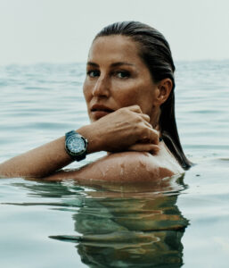 A person with wet hair stands in calm water, their upper body partially submerged. They have a contemplative expression and are resting one arm on the other, showcasing a stylish wristwatch. The serene water reflects their image, creating a tranquil atmosphere. Soft, natural lighting enhances the serene and peaceful ambiance of the scene.