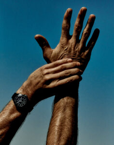 A hand reaches upward against a vibrant blue sky, fingers spread wide. Another hand, adorned with a sleek, dark wristwatch, grasps the extended forearm. The scene conveys a dynamic contrast between the skin tones and the deep sky, highlighting the texture and details of the skin and watch, with sunlight accentuating the highlights and shadows.