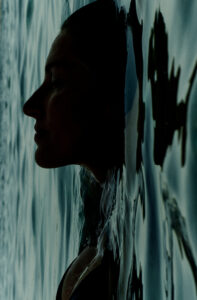 A serene moment captures a person's profile immersed in water, creating a symmetrical reflection. The calm surface gently contours the face and shoulders, blending seamlessly into the dark, shadowy hues. The lighting is soft, emphasizing tranquility and introspection, while ripples give a sense of subtle movement, enhancing the meditative quality of the scene.