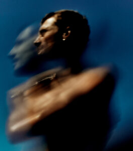 A person is captured against a deep blue background with a soft, blurred effect creating a sense of motion. The subject's face is slightly turned, and their body appears doubled, giving an abstract, ethereal quality. The use of shadows and highlights enhances the mysterious atmosphere, blending colors smoothly to create a dreamlike visual experience.