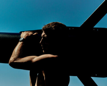 A shirtless man with a sturdy build is silhouetted against a clear blue sky. His muscular arm rests on a large, dark cylindrical object. The sunlight highlights the texture of his skin, and he wears a wristwatch. The strong contrast between light and shadow enhances the scene’s dramatic effect, with minimalistic and rugged vibes.