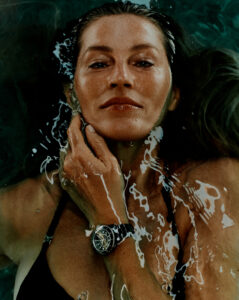 A person with wet hair gracefully lies in water, eyes calm and serene. They wear a black bikini and a sleek watch on their wrist. Water ripples across their face and body, creating elegant patterns. Their hand is gently placed near their chin, with droplets accentuating the natural beauty and peaceful expression captured in this aquatic setting.