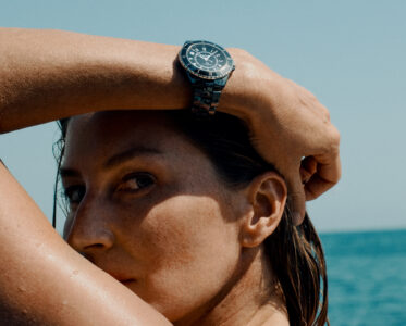 A person is gazing forward while holding an arm above their head. They're wearing a stainless steel watch with a black dial. Their skin glistens slightly, suggesting proximity to water. The background features a clear blue sky meeting the ocean, conveying a sense of tranquility and leisure. Droplets on the skin hint at recent contact with the sea.