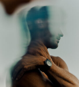 A contemplative figure with a toned physique is seen with a watch on their wrist, hand resting on their shoulder. The scene is softened by a blurred overlay, creating a dreamy, introspective mood. Light reflections add a sense of mystery, while the muted color palette enhances the serene and reflective atmosphere, inviting thoughtful contemplation.