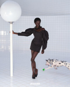 In a minimalist, tiled space, a woman stands elegantly in a dark, puff-sleeved dress, leaning against a tall, spherical light fixture. Her poised demeanor contrasts with a lively, black and white spotted dog, who energetically plays with a small green ball nearby. The scene exudes a blend of sophistication and playful energy within a bright, modern setting.