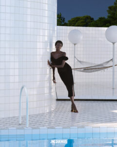 A woman in a chic black dress leans gracefully against a tiled wall beside a swimming pool. The scene features a minimalist aesthetic with white tiles and a clear blue sky. A hammock is in the background, and spherical lamps add a modern touch. The setting exudes elegance and tranquility, blending fashion with architectural design.