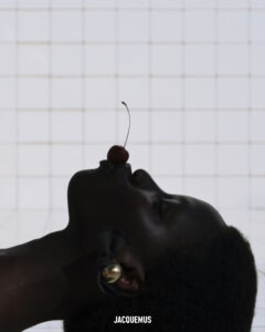 A silhouette of a person's profile is seen against a softly lit grid background. The individual holds a cherry delicately between their lips, its thin stem curving elegantly. Light reflects subtly off the large, shiny earring they wear. The contrast between the dark contour and the bright backdrop creates a striking and artistic composition.