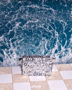 A textured bag, covered in what appears to be tiny crystals or beads, is placed on a tiled surface. The background features a dynamic swirling pattern of white and blue, resembling ocean waves or turbulent water. The scene blends elegance with natural elements, creating a striking visual contrast between the shimmering accessory and the flowing backdrop.