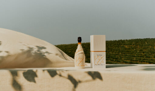 A tranquil scene features a wrapped bottle beside a tall, matching rectangular box. They rest on a beige surface, casting subtle shadows. The background reveals a lush, green field under a clear sky. The composition exudes sophistication and simplicity, accentuated by soft light and shadow play, evoking a serene, upscale atmosphere.