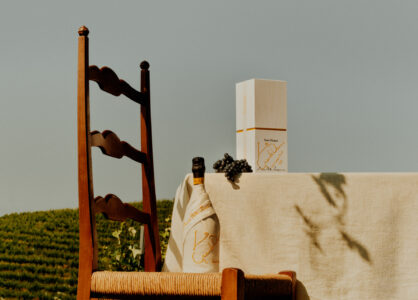 A rustic, wooden chair is positioned beside a table draped in neutral fabric. A bottle wrapped in a cloth and a cluster of dark grapes rest next to an elegant white box. The scene is set outdoors, with lush green vineyards in the background under a clear blue sky, evoking a sense of tranquility and refinement.