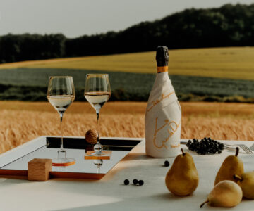 A serene scene features a table set elegantly with two wine glasses on a reflective tray, a wrapped bottle with golden text, and a small wooden block. Black grapes and ripe pears decorate the table. A lush backdrop of golden fields and dense green forest completes the tranquil, picturesque setting under a clear sky.